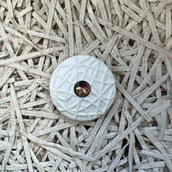 Witte ronde knop met een gematteerde afwerking en een glanzende roestkleurige steen in het midden, geplaatst op een gestructureerde ondergrond.