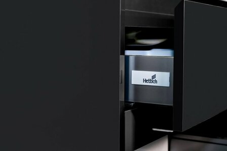 Zwarte lade met een metalen Hettich logo, gedeeltelijk geopend, toont een glanzende binnenkant en eenvoudige, moderne lijnen.