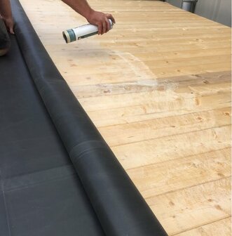 Een man spuit een zwart rubberachtig materiaal op een houten ondergrond met zichtbare nerven.