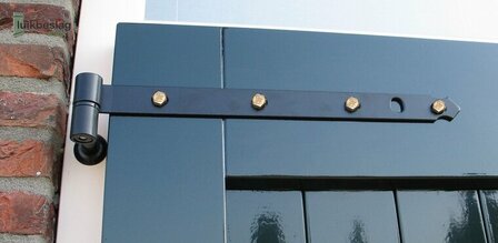 Zwart metalen luikbeslag met gouden schroeven, bevestigd aan een groene deur met een bakstenen muur op de achtergrond.