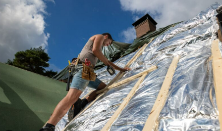 Een man in een tanktop en korte broek werkt op een hellend dak bedekt met een reflecterende folie, onder een blauwe lucht.