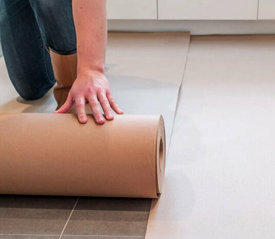 Een hand rolt een grote rol bruine papieren vilt op een lichte vloer, met jeans en een neutrale achtergrond.
