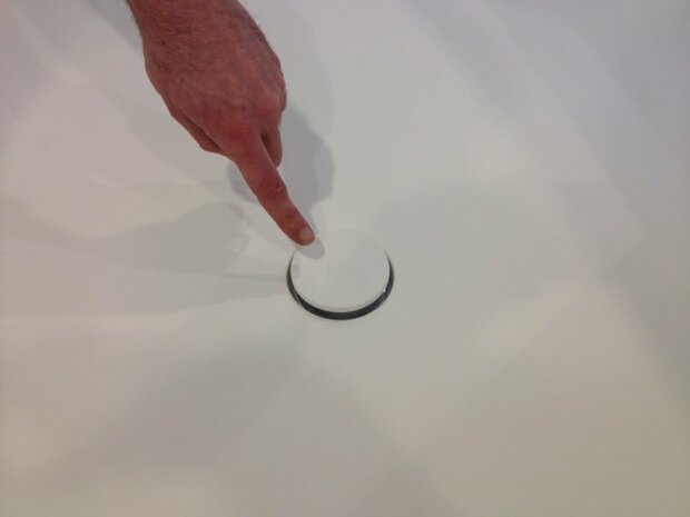 Een hand wijst naar een wit, rond knopje met een zwarte rand op een gladde, witte ondergrond.