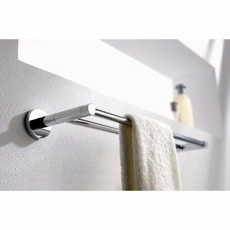 Chromen handdoekhouder bevestigd aan een witte muur, met een hangende, witte handdoek en een flesje shampoo op de achtergrond.