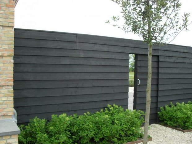 Zwarte houten schutting met verticale planken, een open deur aan de rechterkant en groene struiken aan de onderkant.