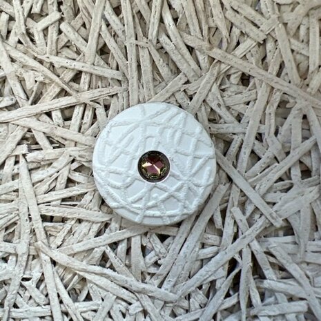 Witte ronde knop met een gematteerde afwerking en een glanzende roestkleurige steen in het midden, geplaatst op een gestructureerde ondergrond.