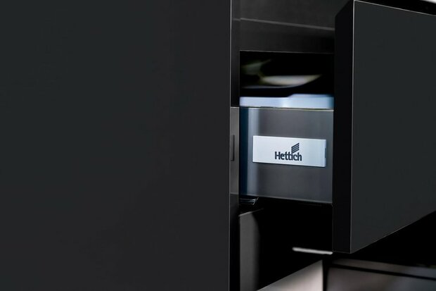 Zwarte lade met een metalen Hettich logo, gedeeltelijk geopend, toont een glanzende binnenkant en eenvoudige, moderne lijnen.