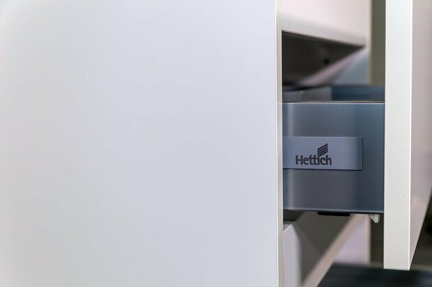 Een lichte kast met een subtiele metalen lade, voorzien van een Hettich logo op een grijze ladefront.