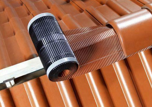 Een zwart rolmechanisme ligt op een glanzende, terracotta kleurige dakpan, met een metalen leuning ernaast.