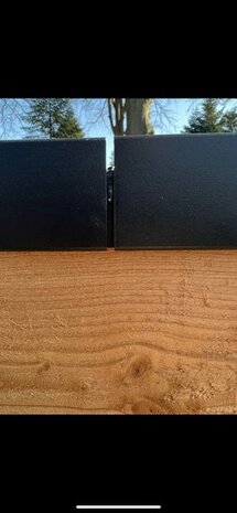 Zwarte metalen panelen met een matte afwerking, geplaatst boven een ruwe houten ondergrond. In de achtergrond bomen en blauwe lucht.