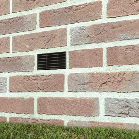 Een zwart ventilatierooster tussen lichtroze met witte bakstenen, met een groene graslijn aan de onderkant.