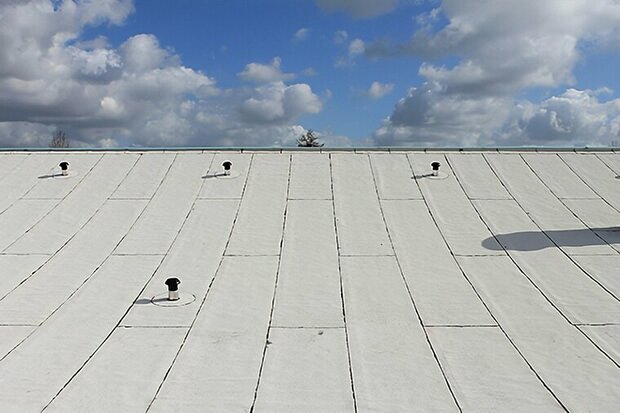 Een lichtgrijze, hellende dakbedekking met meerdere zwarte ventilatie-openingen onder een blauwe lucht vol witte wolken.