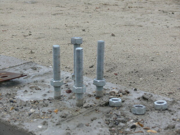 Drie verzinkte bouten met moeren, naast een paar losse moeren, op een zanderige ondergrond.